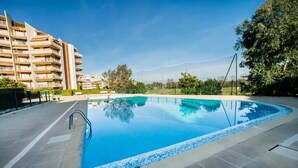 Uma piscina com borda revestida, rodeada por um espaço pavimentado e um edifício com varandas.