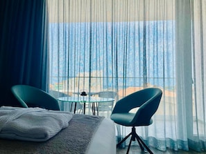 A hotel room with a bed, two chairs, a small table, and a view of the sea through sheer curtains.