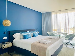 A hotel room with a large bed, a bedside table with a phone, a chair, a small table, and a view of the outdoors.