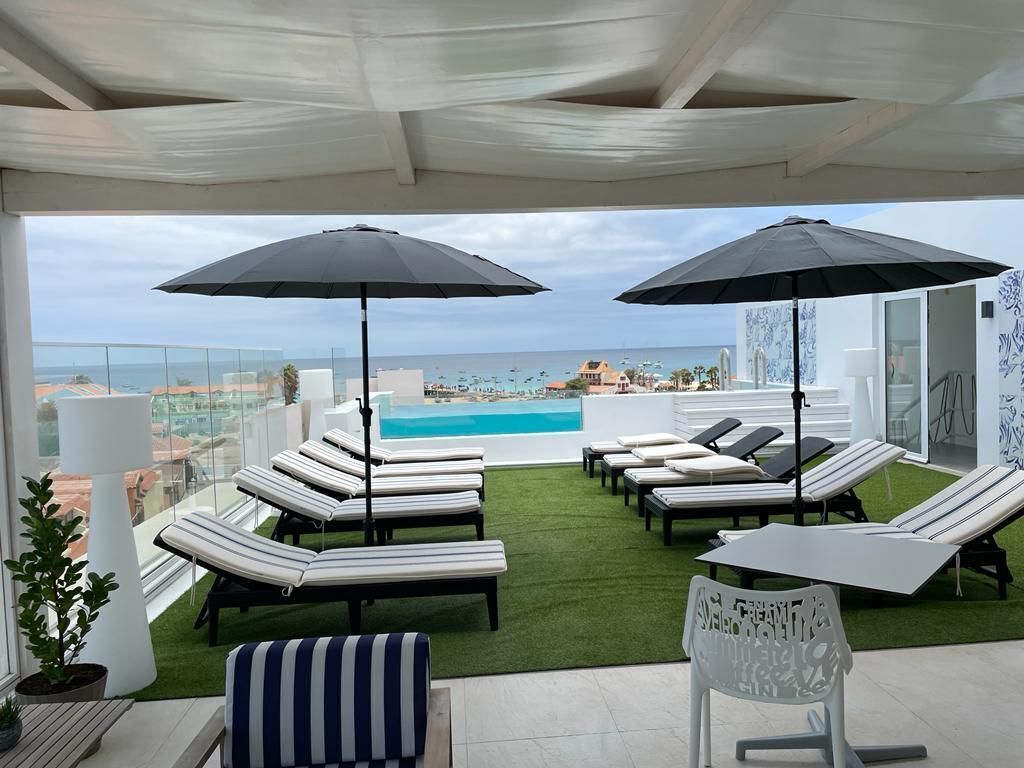 A rooftop lounge area with sun loungers and umbrellas, overlooking a pool and cityscape.