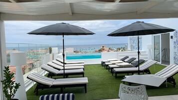 A rooftop lounge area with sun loungers and umbrellas, overlooking a pool and cityscape.
