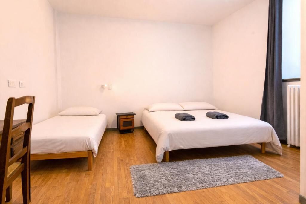A small, single-room accommodation with two beds, a wooden chair, a small bedside table, and a grey rug.