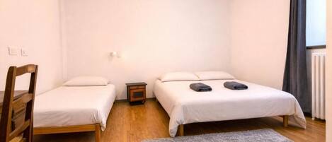 A small, single-room accommodation with two beds, a wooden chair, a small bedside table, and a grey rug.