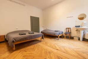 Ein Zimmer mit zwei Betten, einem Holzboden im Fischgrätmuster, einer grünen Tür, einem kleinen Holzstuhl und einem weißen Waschbecken mit Spiegel.