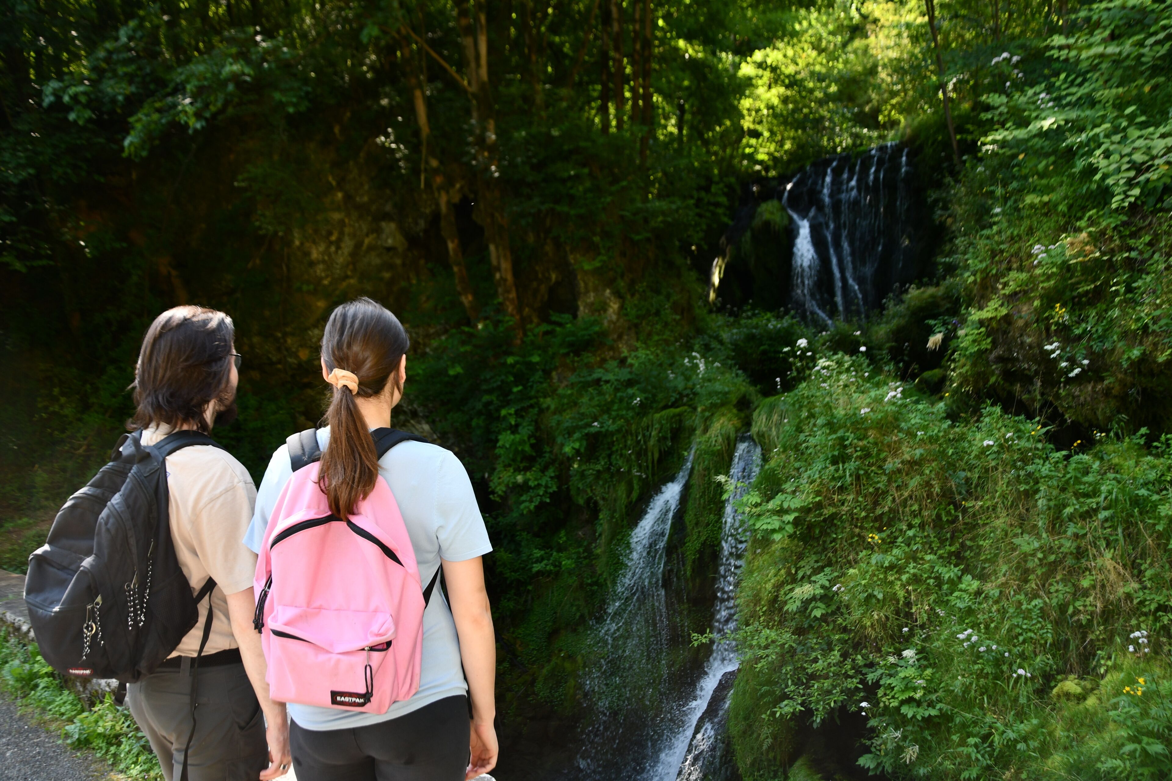 Twee mensen wandelen langs een pad naast een waterval in een bebost gebied.