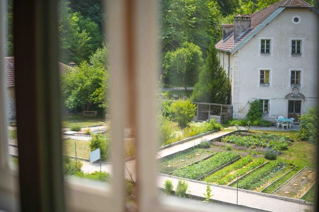 Blick durch ein Fenster auf ein Gebäude mit Garten, hellem Außenbau und einer sichtbaren Tür.