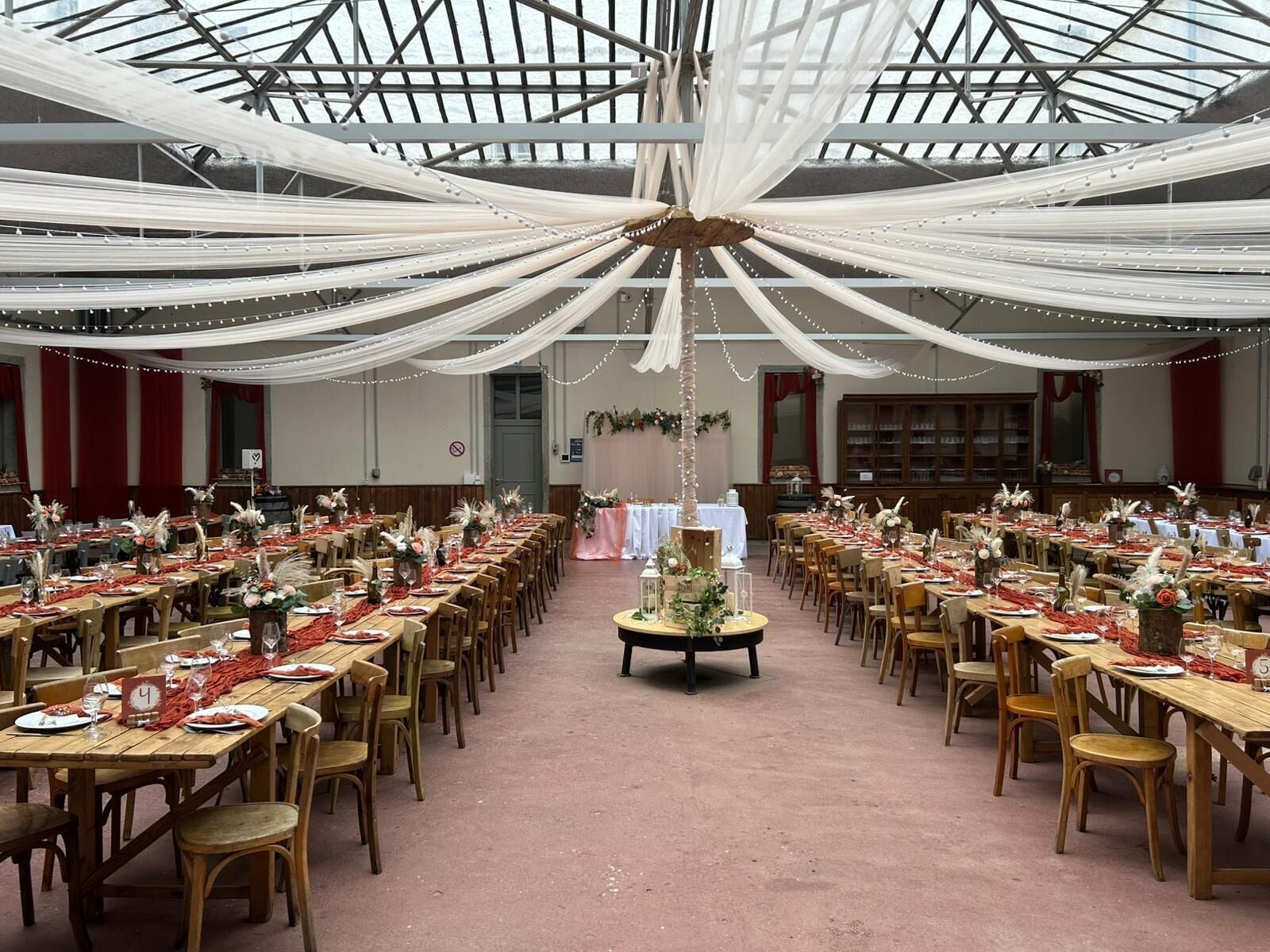 A large hall with tables set for dining, a central decorative piece, and a high ceiling with draped fabric.