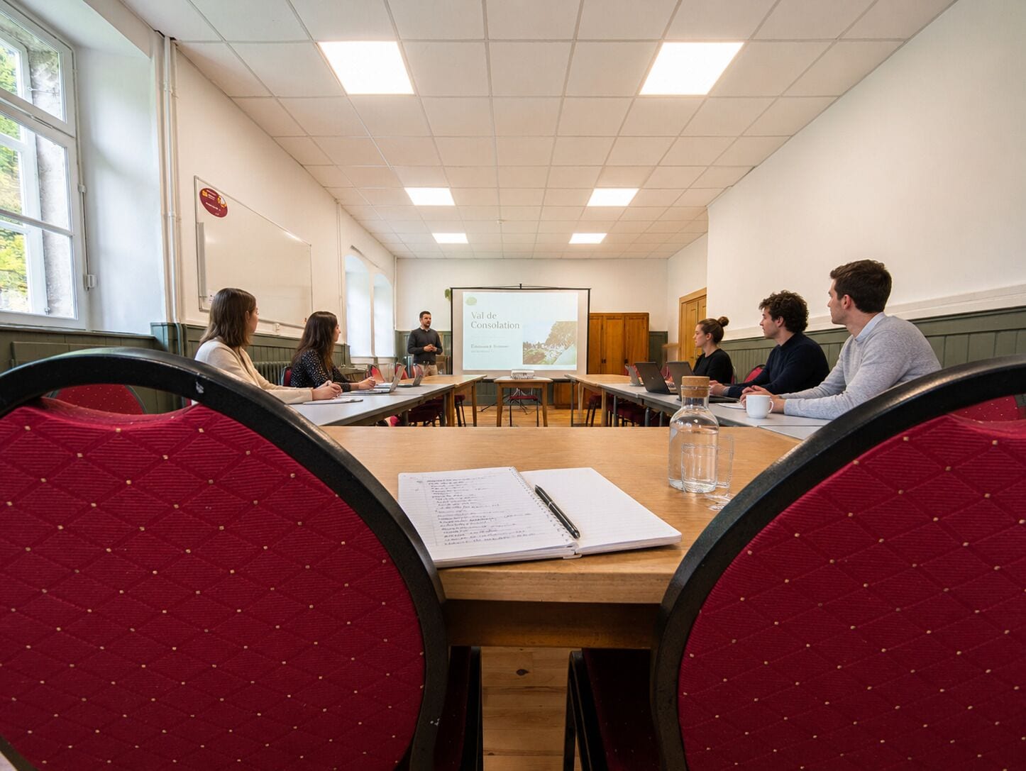A conference room with a presentation screen, wooden tables, and red upholstered chairs.