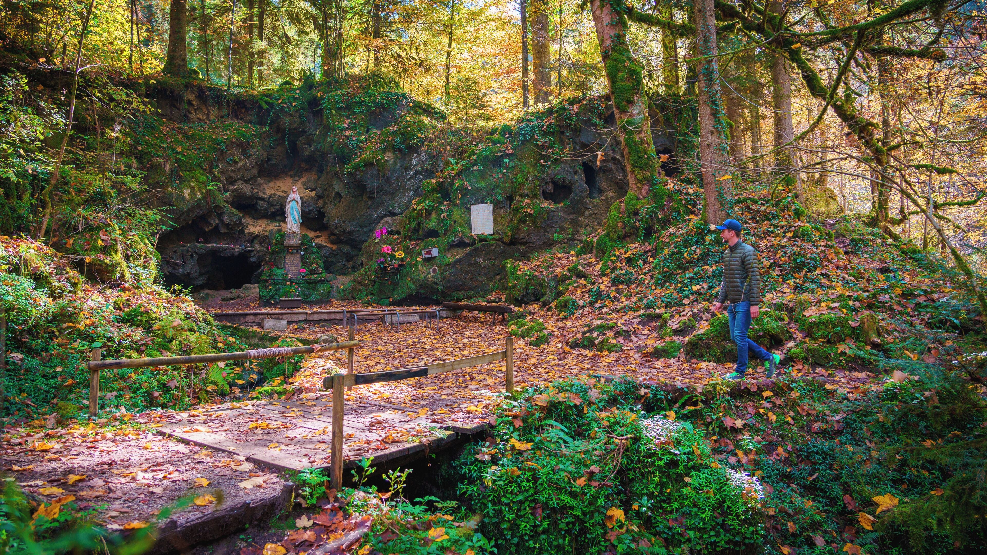 A forested area with a wooden bridge, a statue, and a person walking.