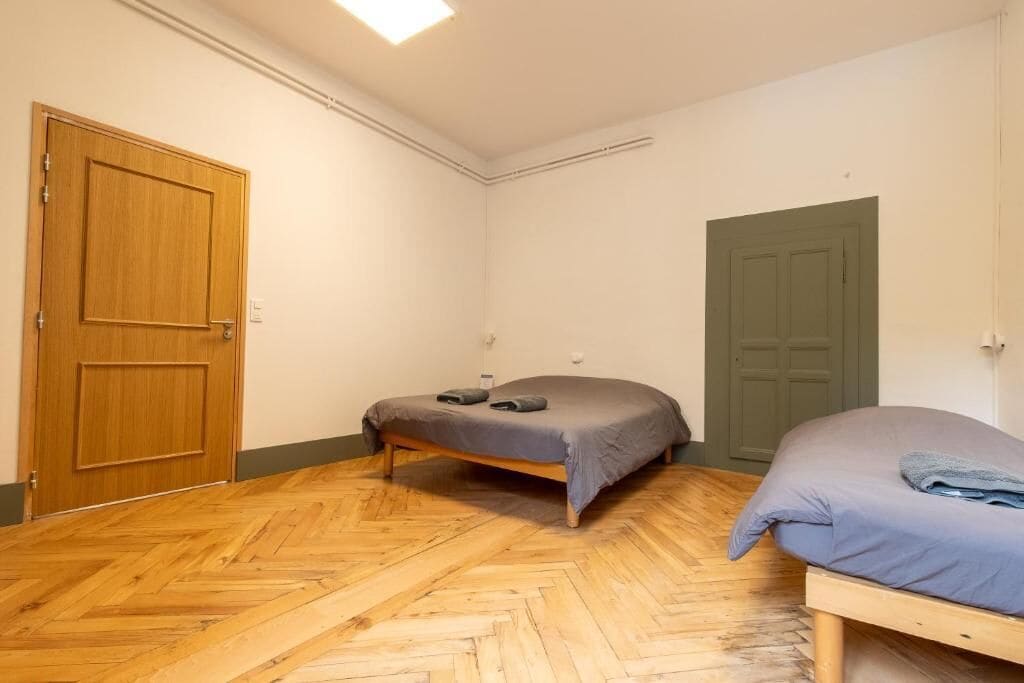 A room with a herringbone wooden floor, two beds with grey bedding, a wooden door, and a green door.