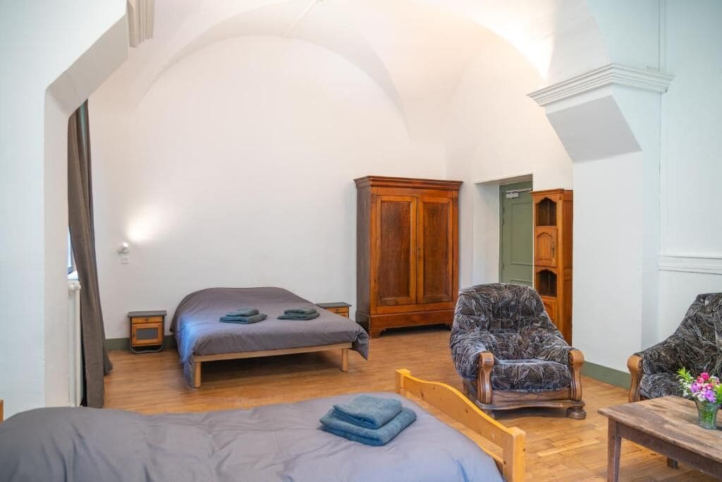 A bedroom with a bed, two chairs, a wooden wardrobe, and a small table with a vase.