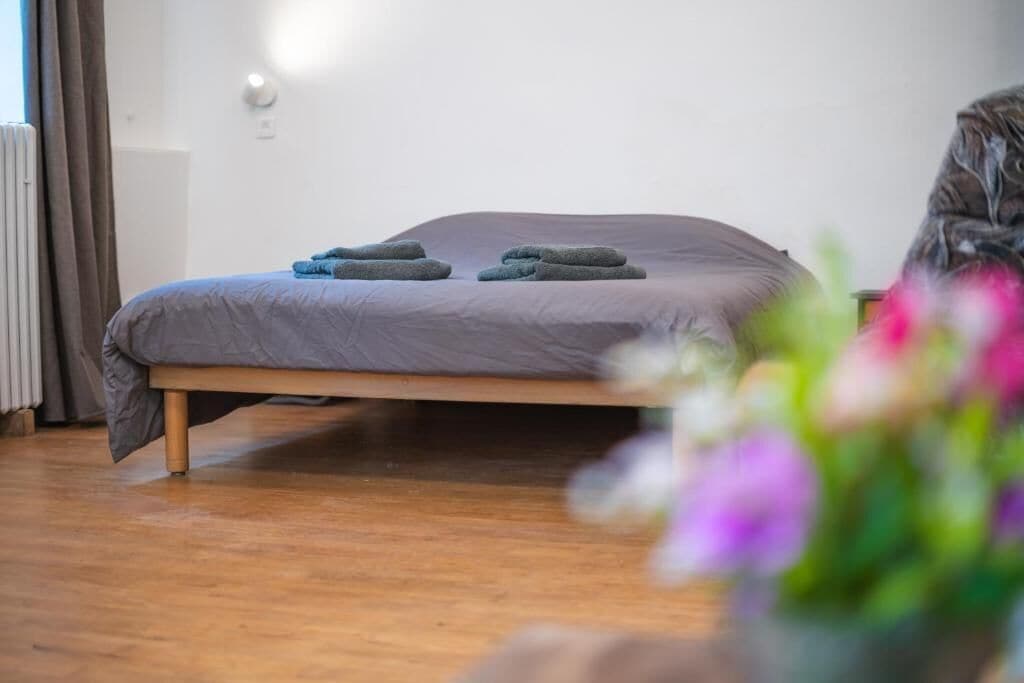 Un lit simple avec un matelas gris et un cadre en bois, un radiateur et une fenêtre avec des rideaux.