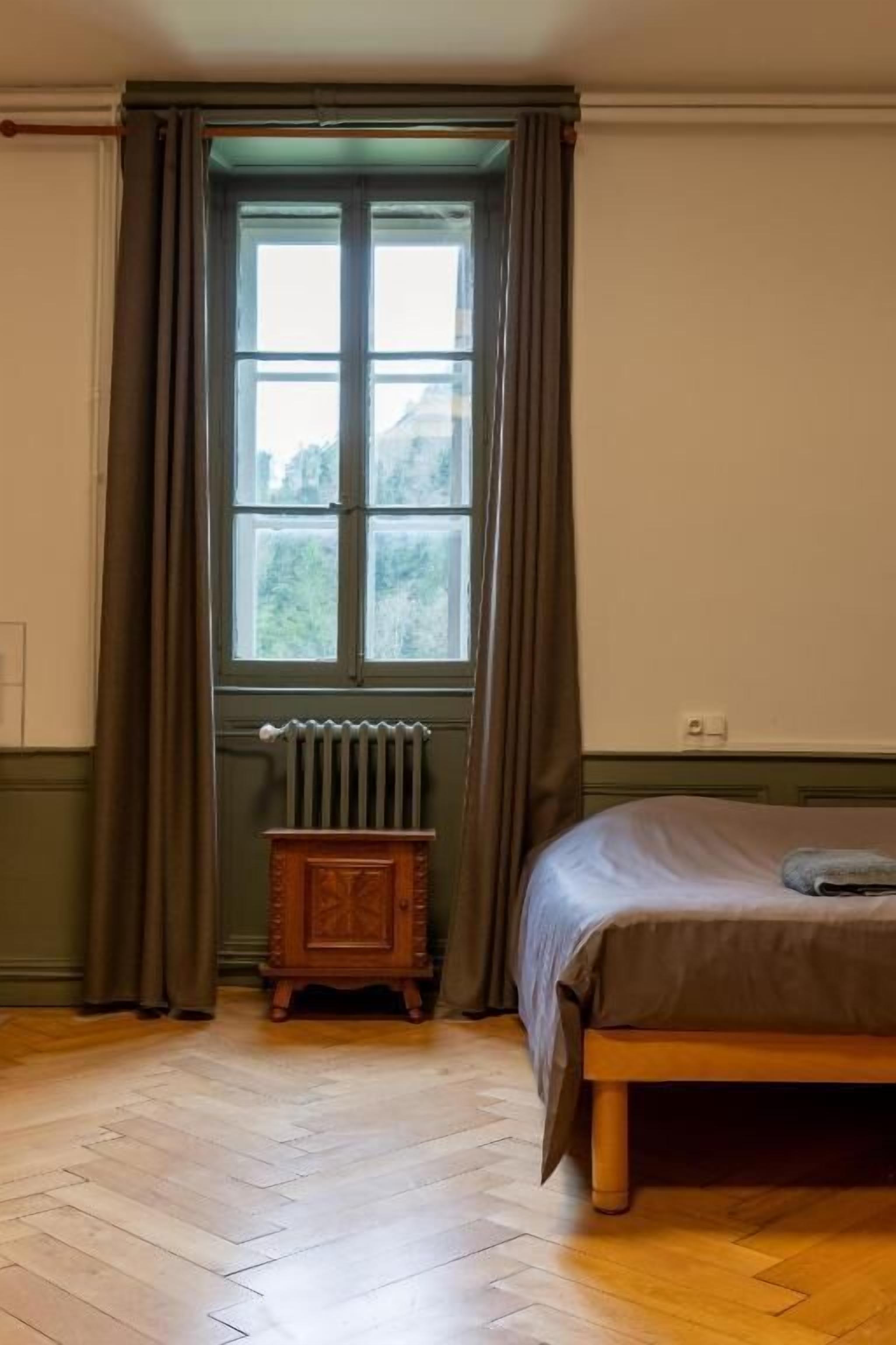 Ein Zimmer mit einem Bett, einem Heizkörper, einem Fenster mit Vorhängen und einem Holzschrank.