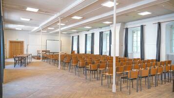 Een grote zaal met rijen stoelen en een lange tafel, witte muren en grote ramen.