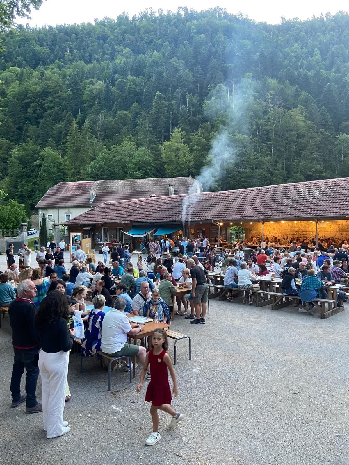 Un espace extérieur animé, aménagé pour les repas, avec des tables et des bancs, entouré d’une colline boisée.