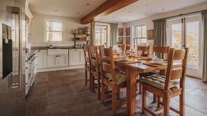 A spacious kitchen with white cabinets, a wooden dining table set for six, and a stone-tiled floor.