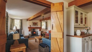 A cozy living room with a fireplace, wooden beams, a sofa, a table, and a kitchen area with cabinets.