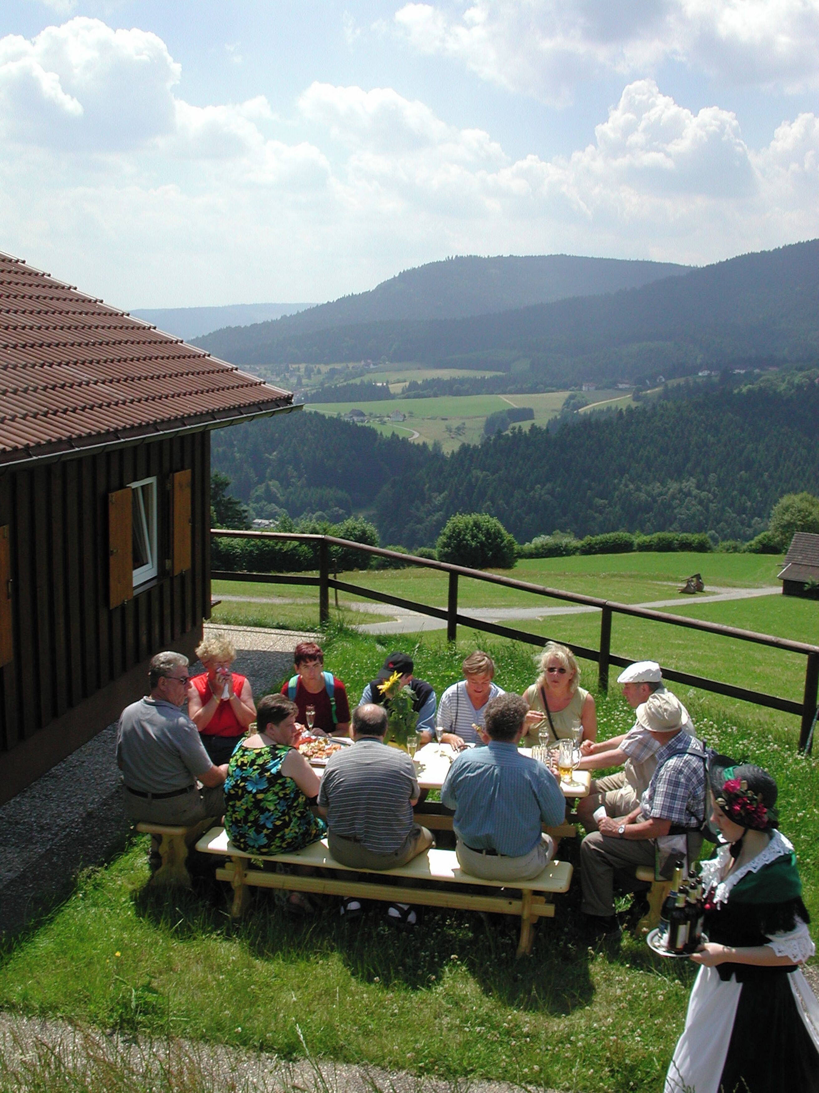 Eine Gruppe von Menschen speist draußen und genießt dabei die Aussicht auf Hügel und ein Holzgebäude im Hintergrund.