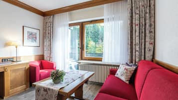 Ein gemütliches Zimmer mit einem roten Sofa, einem Holztisch mit einer Pflanze, einem Schreibtisch mit einer Lampe und einem Fenster mit Vorhängen.