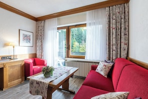A cozy room with a red sofa, a wooden table with a plant, a desk with a lamp, and a window with curtains. - Sackmann Landhaus (Baiersbronn)