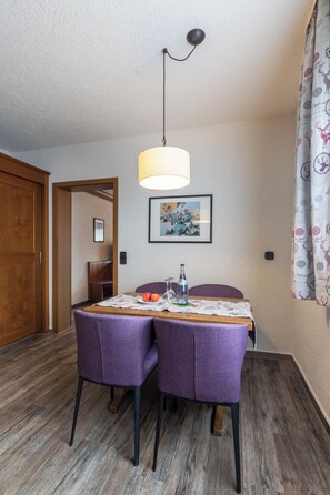 Un comedor con una mesa preparada para dos personas, dos sillas moradas, una lámpara colgante, un cuadro en la pared y una puerta que da a otra habitación.