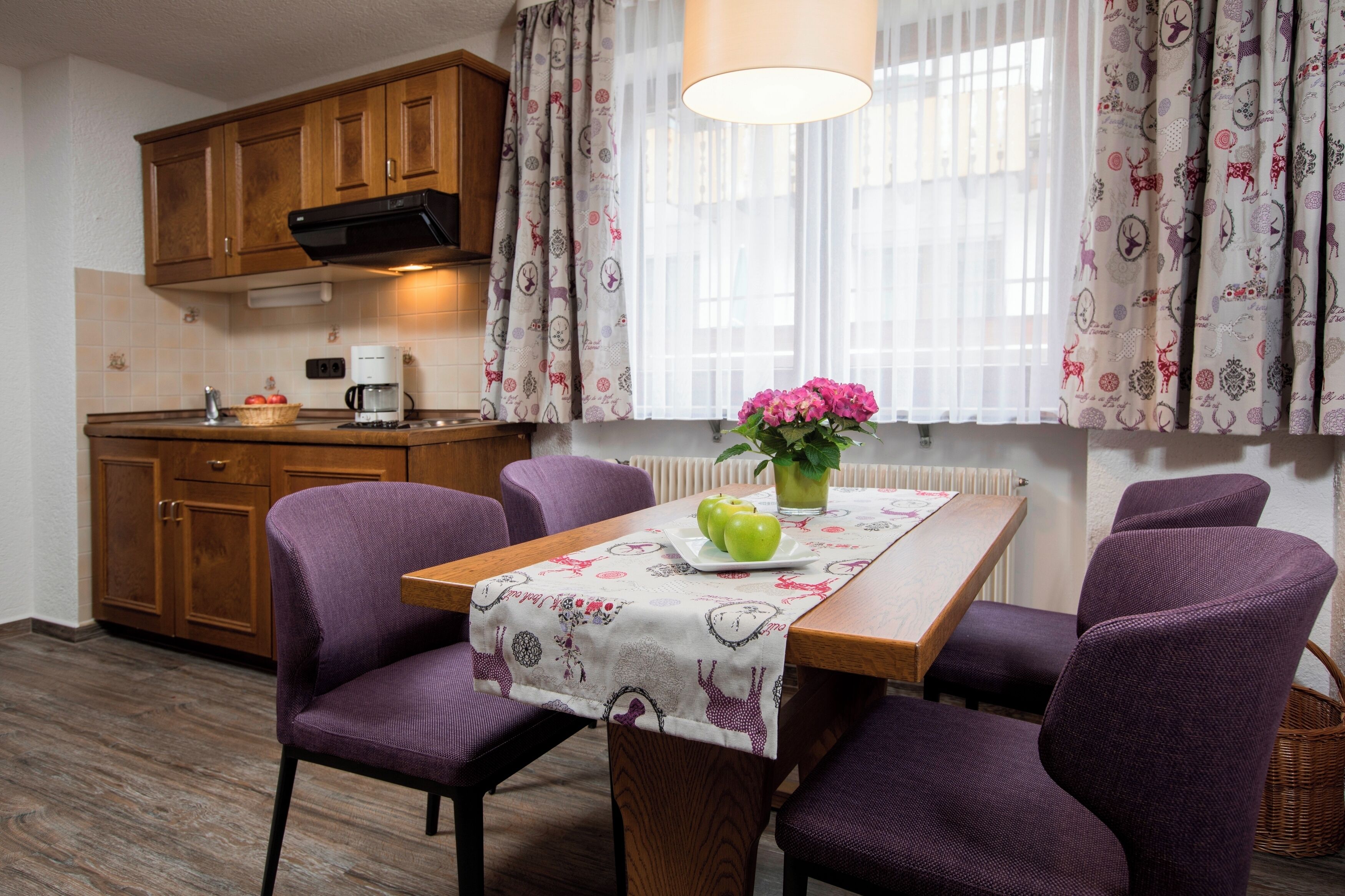 Un comedor con una mesa lista para dos personas, sillas moradas, una cafetera y una ventana con cortinas estampadas.