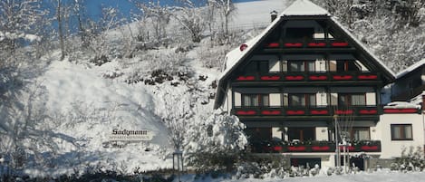 Un bâtiment de style chalet traditionnel, avec un toit enneigé, entouré d’un paysage hivernal et d’arbres couverts de neige.