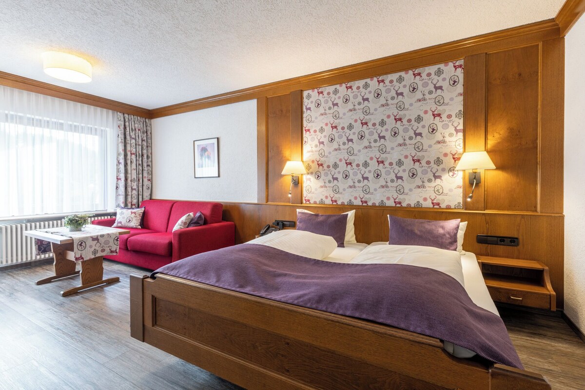 A hotel room with a wooden bed, a red sofa, a small table with a lamp, and a patterned wallpaper.