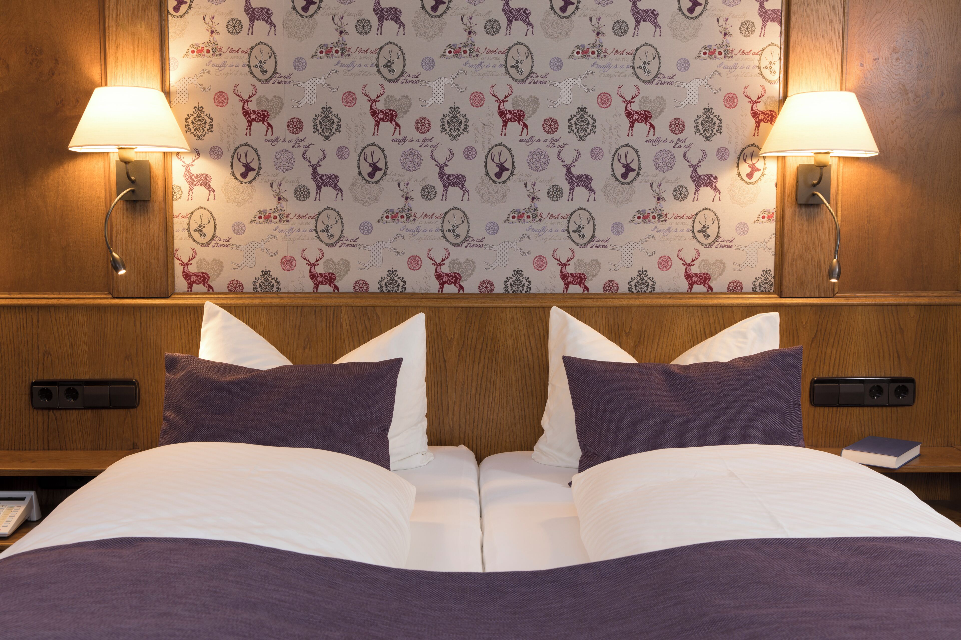 Una habitación de hotel con cabecera de madera, dos lámparas de noche, un papel tapiz estampado con ciervos y corazones, y una cama bien hecha con ropa de cama blanca y morada.