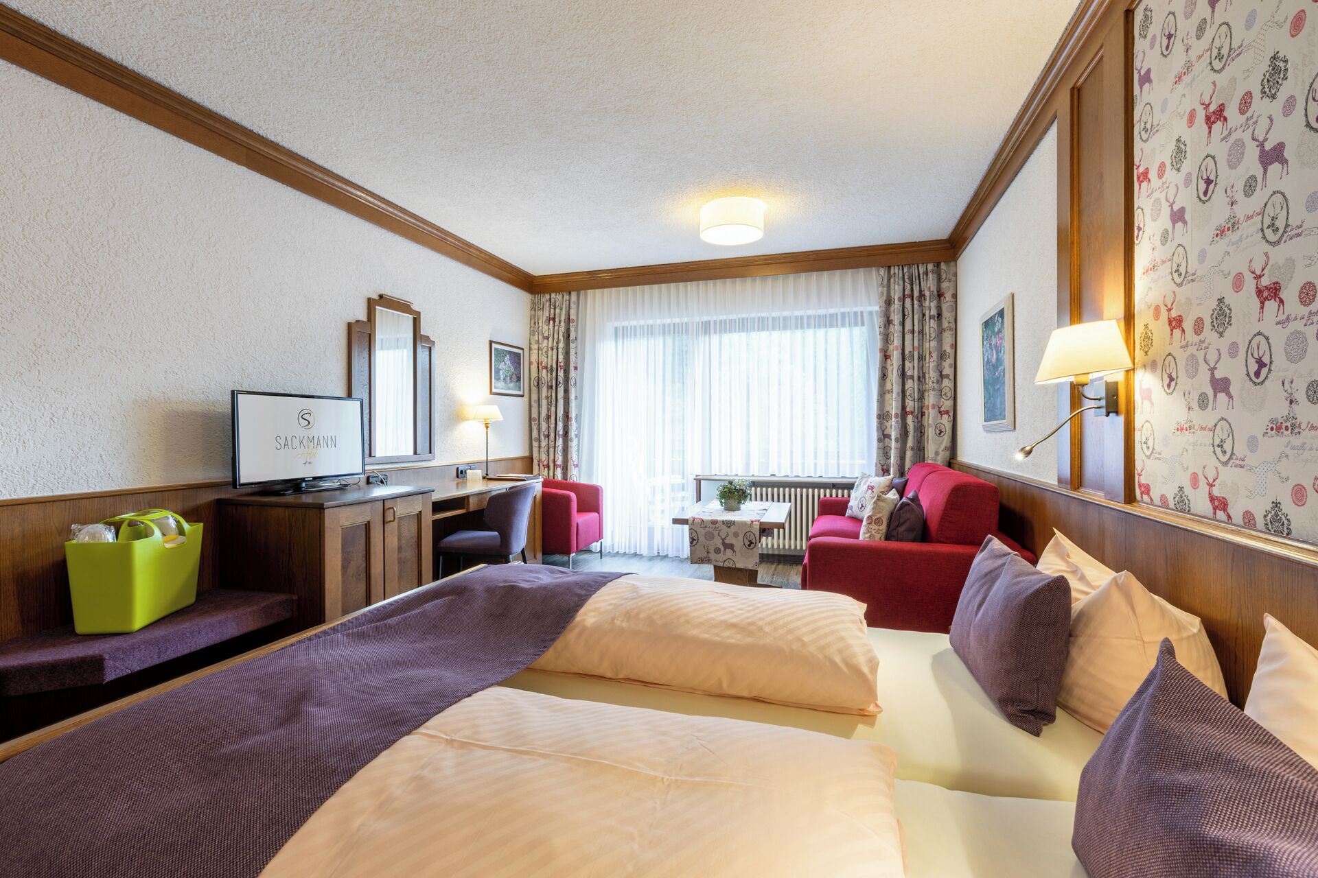 A hotel room with a bed, two chairs, a desk, a television, and a window with curtains.