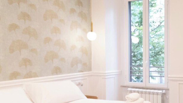 Una cama bien tendida con toallas dobladas, una pared con un diseño de hojas, una ventana con vistas a zonas verdes y un radiador.