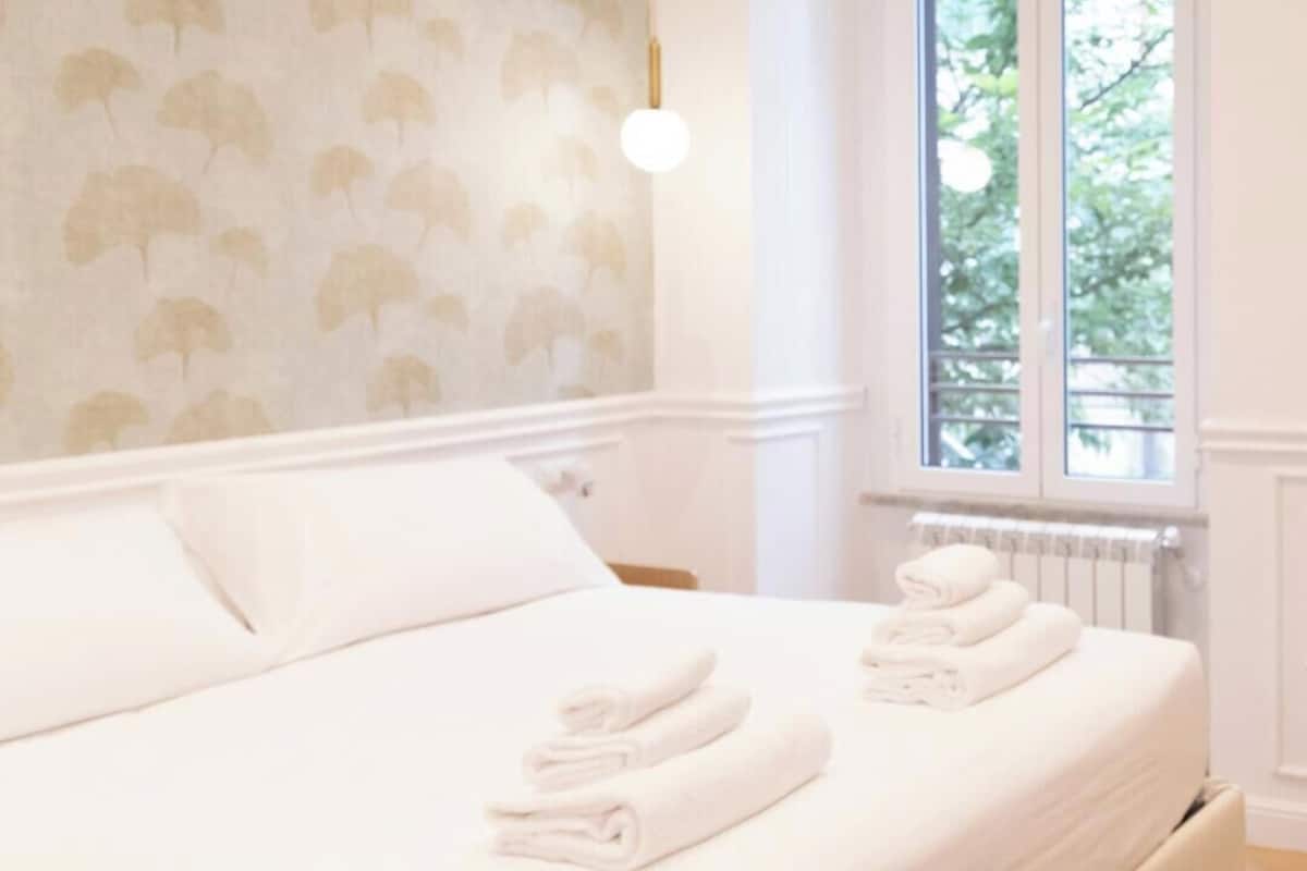 Una cama bien tendida con toallas dobladas, una pared con un diseño de hojas, una ventana con vistas a zonas verdes y un radiador.
