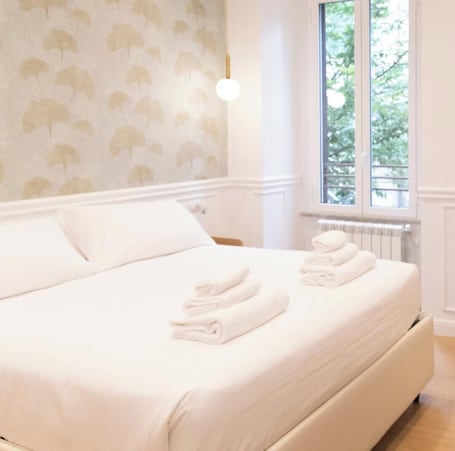 A neatly made bed with folded towels, a wall with a leaf pattern, a window with a view of greenery, and a radiator.