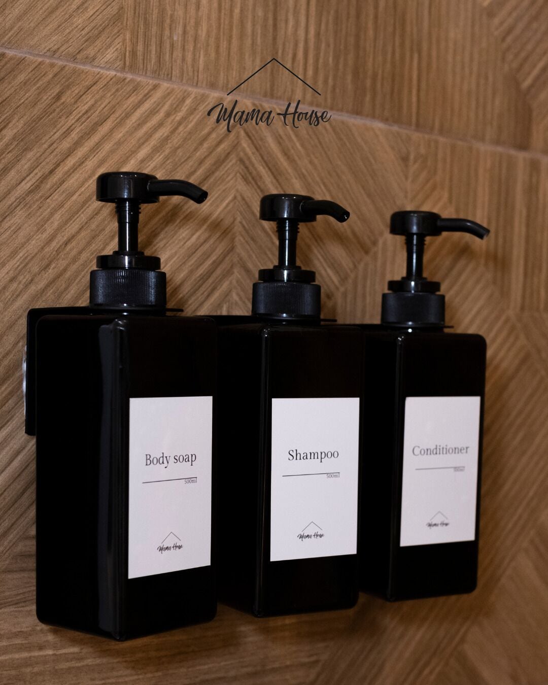 Trois flacons noirs équipés de pompes, étiquetés « savon pour le corps », « shampoing » et « après-shampoing », chacun ayant une étiquette blanche et une pompe noire.