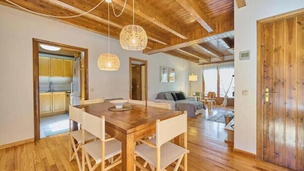 Uma sala de jantar com mesa e cadeiras de madeira, cozinha ao fundo e área de estar com sofá e abajur.