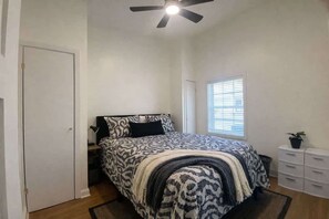 Un dormitorio con cama, ventilador de techo, ventana con persianas, mesita de noche y cómoda.