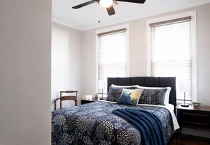 Un dormitorio con cama, dos ventanas con persianas, ventilador de techo, mesita de noche y una pequeña mesa.