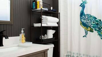 Une salle de bain avec des toilettes blanches, un meuble-lavabo en bois, une étagère contenant des articles de toilette et un rideau de douche orné de motifs de paon.