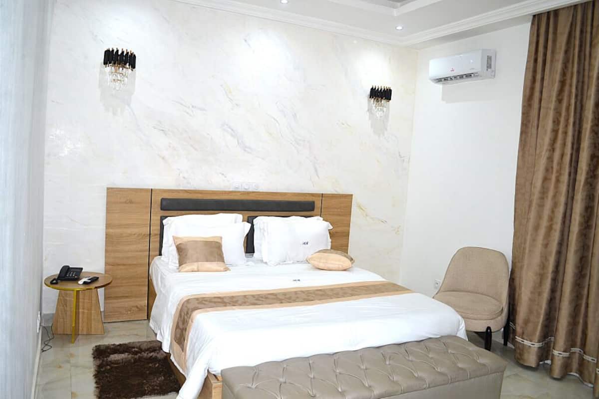 A hotel room with a bed, bedside table, chair, and an air conditioning unit.