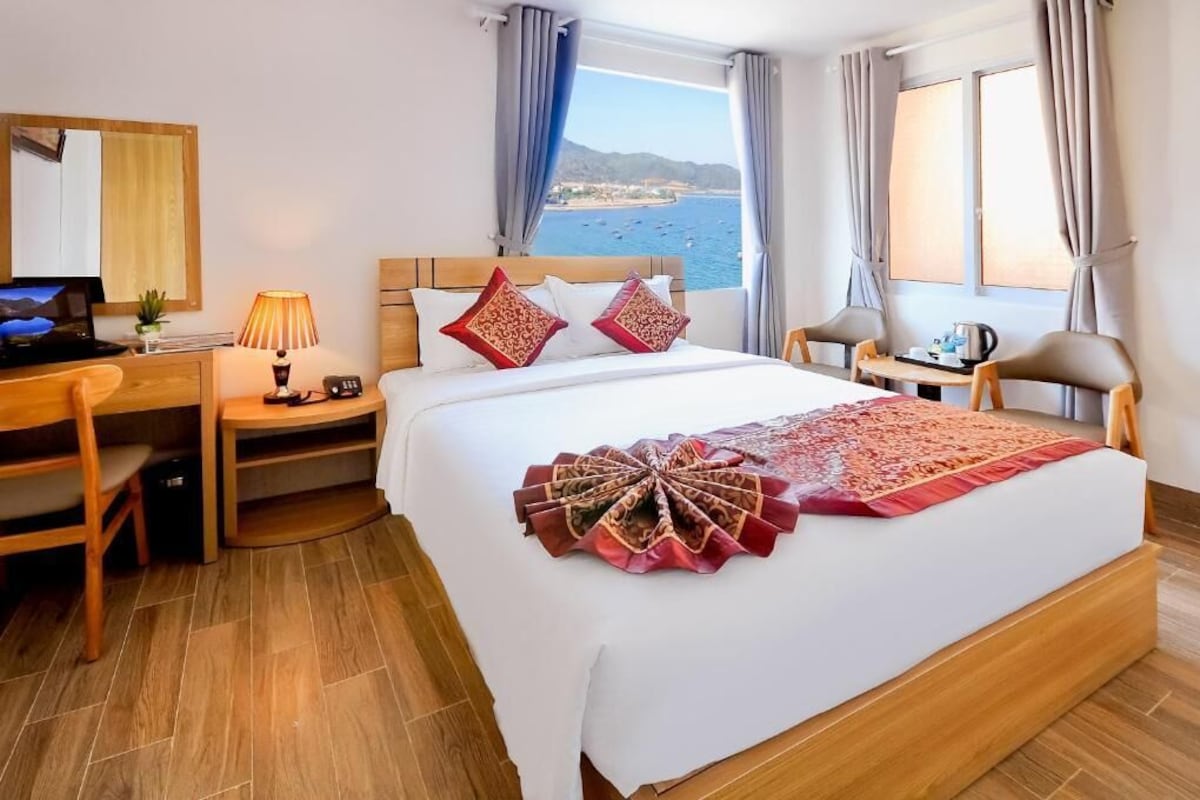Quarto de hotel com cama, escrivaninha, cadeira, luminária e vista para o mar.