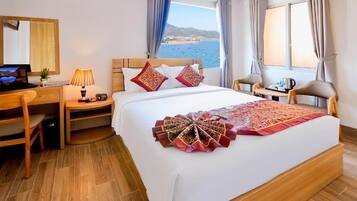 A hotel room with a bed, a desk, a chair, a lamp, and a view of the sea.