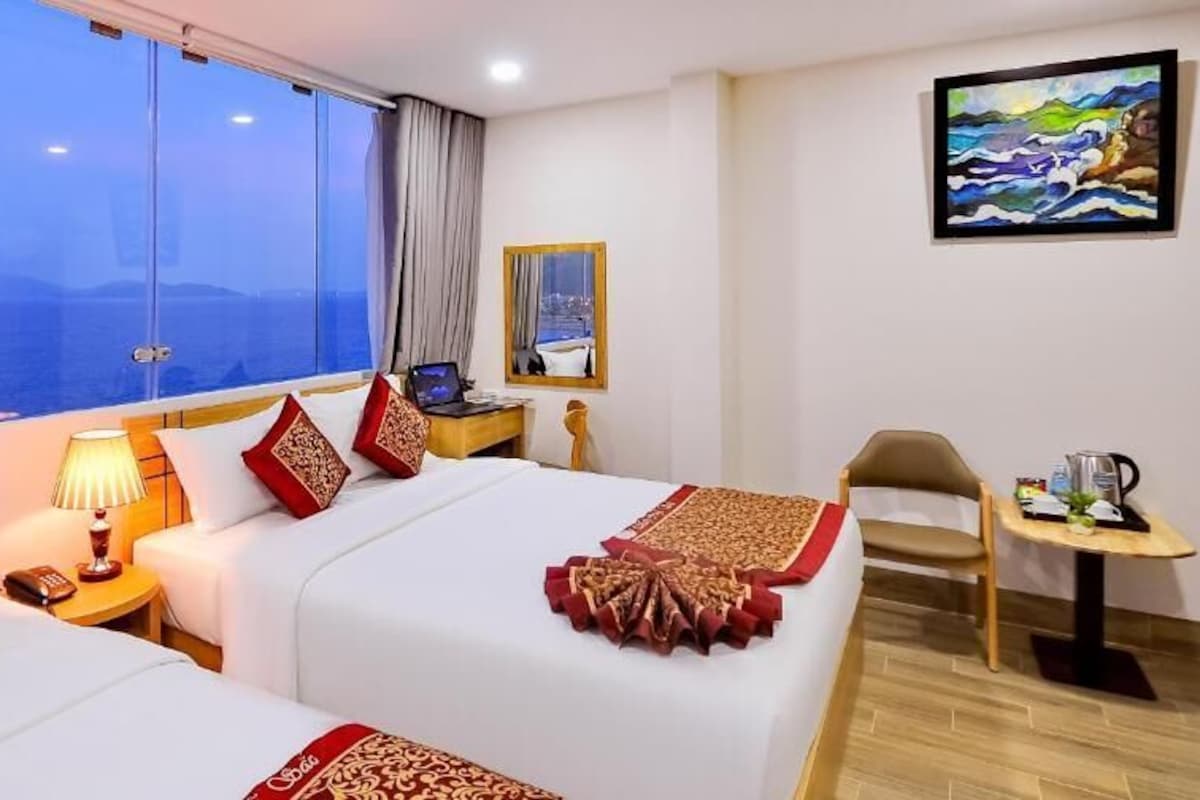 Quarto de hotel com duas camas, uma escrivaninha, uma cadeira, um quadro e vista para o mar.