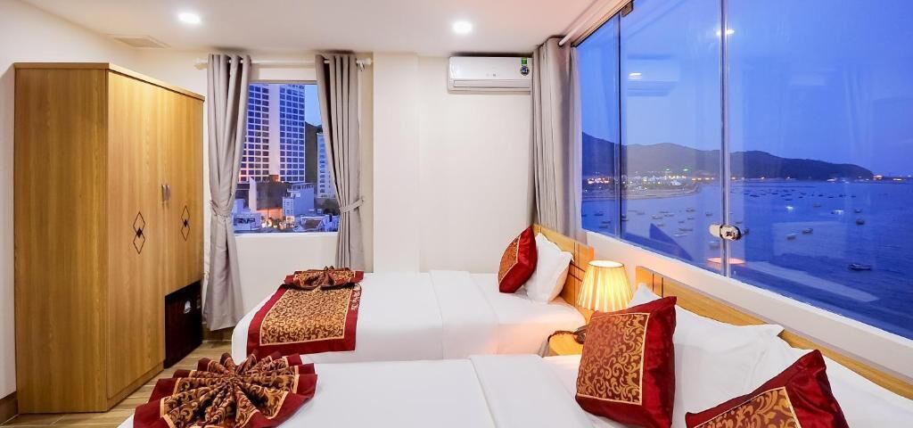 Quarto de hotel com duas camas, um guarda-roupa, ar condicionado e uma janela ampla com vista para a cidade e o mar.