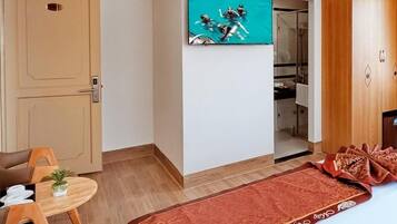 A hotel room with a TV mounted on the wall, a wooden wardrobe, a small table with a plant, and a red patterned rug.