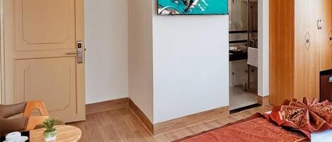 Habitación de hotel con televisor montado en la pared, armario de madera, una mesita con una planta y una alfombra roja estampada.