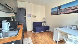 Ein kompakter Wohnraum mit Küchenzeile, einer Couch, einem Schreibtisch und einem Bild von einer Strandansicht.