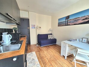 A compact living space with a kitchenette, a sofa, a desk, and a painting of a beach scene. - Studio Cosy Mer & Bus St-Malo (Dinard)