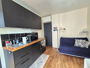 A compact kitchen with a microwave, kettle, and sink, a dark sofa with patterned cushions, and a small plant on the counter. - Studio Cosy Mer & Bus St-Malo (Dinard)