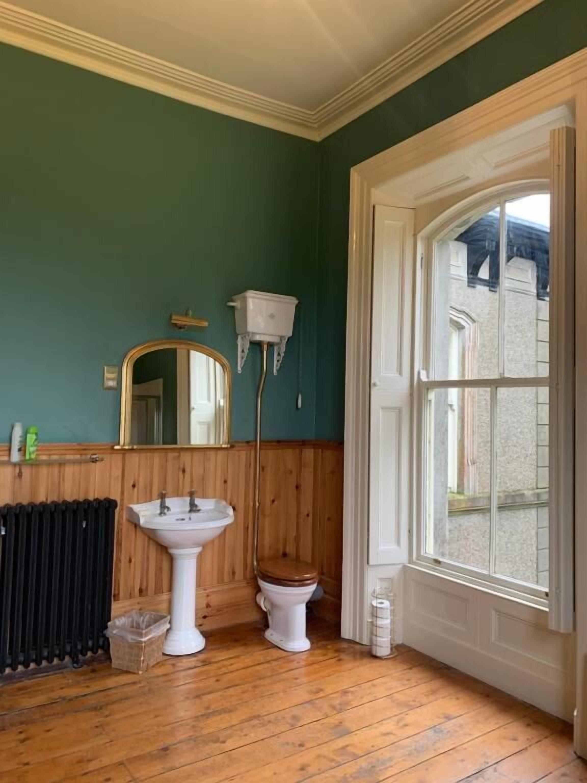 Un baño con piso de madera, un lavamanos blanco, inodoro, espejo y ventana.