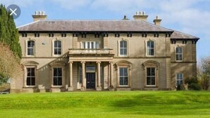 A large, stately building with a symmetrical facade, a central entrance with columns, and a well-maintained lawn in front. - Ballynacree Manor House  (Ballymoney)